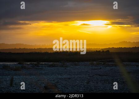 Die Sonne platzt durch Wolken und lässt sich in der Silhouette von Okarito, Neuseeland, in goldenem Farbton über die Landschaft ziehen. Stockfoto