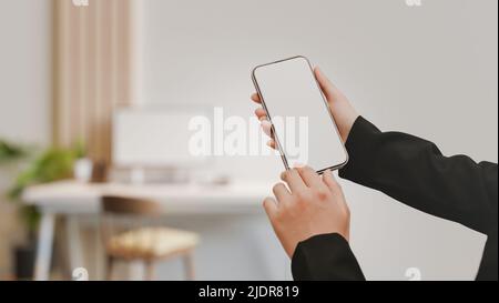Eine Geschäftsfrau hält sich in einem formellen Anzug mit einem Smartphone-Modell mit weißem Bildschirm über einem verschwommenen, minimalweißen Büroarbeitsplatz im Hintergrund. 3D Rendern, Stockfoto