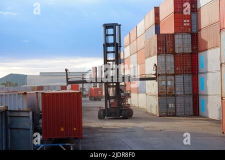 Gabelstapler, die Frachtcontainer im Logistikhof mit Stapeln von Frachtcontainern im Hintergrund abtransportieren. Stockfoto