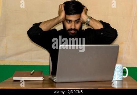Ein junger Inder, der auf einem Stuhl sitzt und gestresst auf den Laptop-Bildschirm blickt. Selektiver Fokus auf das Gesicht. Stockfoto