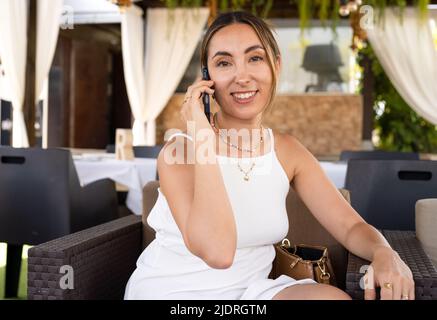 Junge charmante Frau mit Mobiltelefon, während allein im Coffee Shop in der freien Zeit, attraktiven weiblichen mit niedlichen Lächeln in Sprechen Stockfoto