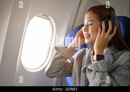 Wunderschöne und schöne asiatische Geschäftsfrau in legerem Anzug, die während des Fluges Musik auf ihren modernen kabellosen Kopfhörern hört. Flugzeugtransport Stockfoto