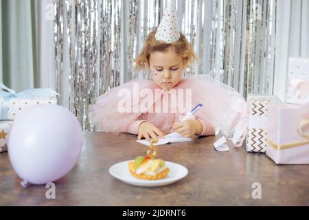 Geburtstag ist fünf Jahre alt. Charmantes Mädchen, festlich gekleidet, umgeben von Luftballons, Geschenken und Kuchen mit der Nummer fünf, schreibt fleißig Dank für Stockfoto