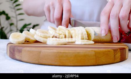 Frau schneidet Banane auf Holzbrett und bereitet Smoothie oder Milchshake zu Stockfoto