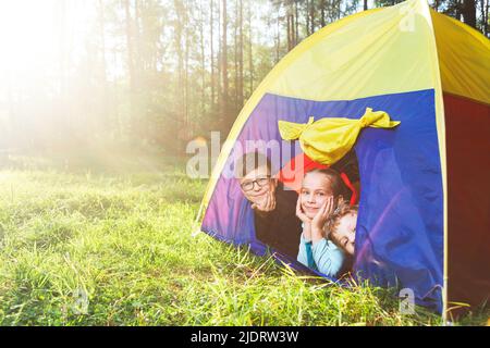 Kinder liegen im Zelt auf einer sonnigen Wiese und genießen sommerlichen Waldcampen. Die Sonne scheint durch die Bäume. Konzept für einen Sommerurlaub mit der Familie Stockfoto
