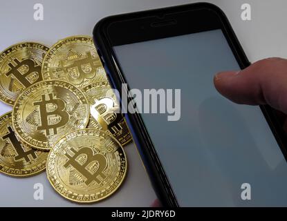 Bitcoin-Münzen aus goldenem Metall mit einer Männerhand neben ihm und einem Smartphone Stockfoto