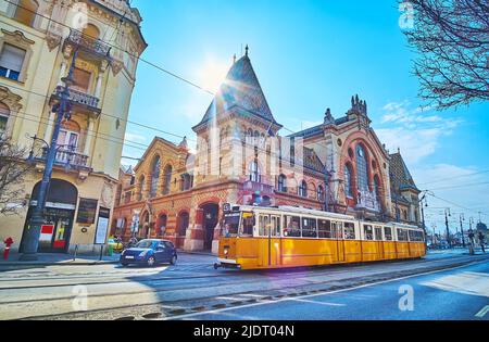 Die urbane Szene mit einer alten gelben Straßenbahn, dem Gebäude der Central Market Hall und der hellen Sonne, die über dem Dach scheint, Budapest, Ungarn Stockfoto