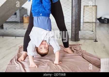 Junge, der mit der Hilfe der Mutter einen Handstand macht, während er zu Hause im Wohnzimmer spielt Stockfoto