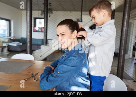 Mutter sitzt am Tisch im Wohnzimmer und lässt ihren Sohn die Haare kämmen Stockfoto