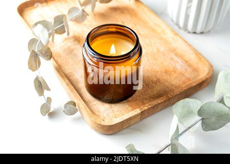 Brennende aromatische Kerze und Eukalyptuszweig auf dem Tisch auf Holztablett. Gemütlicher Lebensstil. Stockfoto