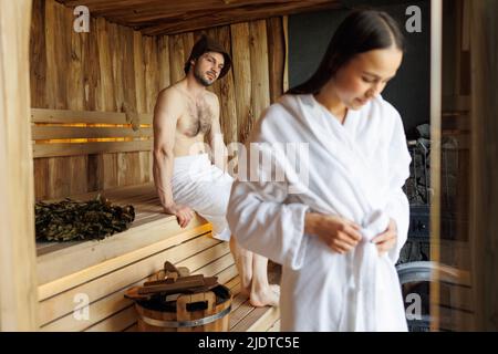 Hübsche junge Frau in der Sauna, die einen Bademantel und ihren Mann im Hintergrund bindet. Paar in der Sauna Stockfoto
