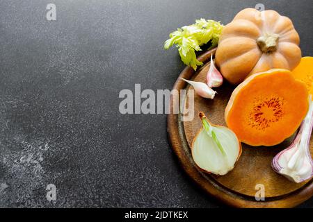 Frisches, helles Gemüse wird auf einem hölzernen Schneidebrett angelegt: butternusskürbis, grüne Sellerie-Blätter. Schwarzer ungleichmäßiger Hintergrund, Kopierbereich. Stockfoto