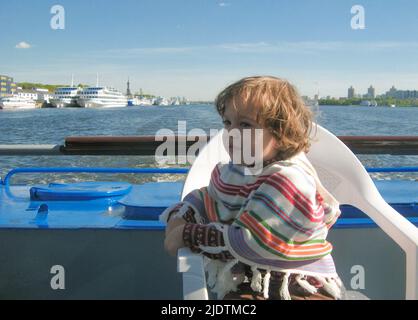 Ein niedliches Kleinkind-Mädchen in einem gestreiften Poncho sitzt an Bord des Schiffes und schaut ans Ufer. Das Boot fährt auf einem breiten Fluss. Das Konzept des Reisens o Stockfoto