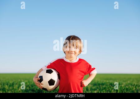 Glückliches Kind gibt vor, ein Fußballspieler zu sein. Porträt eines Kindes im Freien. Junge gegen grünes Feld. Erfolg und Siegerkonzept Stockfoto