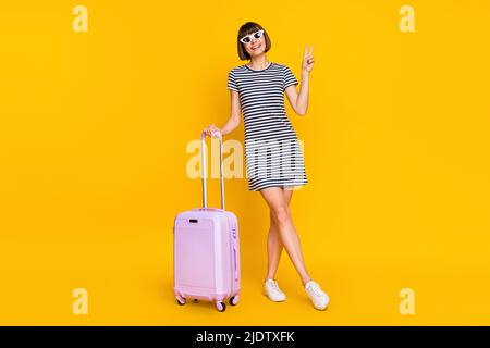 In voller Länge Foto von coolen Brünette junge Dame Stand zeigen V-Zeichen mit Tasche tragen Kleid Turnschuhe Brille isoliert auf gelbem Hintergrund Stockfoto