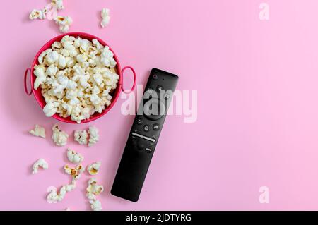 Popcorn in einer Metallschüssel und eine Fernbedienung auf einem rosa Hintergrund mit Platz für Kopien. Das Konzept der Freizeit, Filme, Shows, Sport Stockfoto