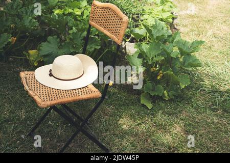 Sommerhut für Frauen auf einem alten Korbstuhl im Garten, ländliches Leben und wachsendes Lebensmittelkonzept Stockfoto