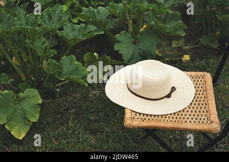 Frauen-Sommerhut auf einem Korbsessel im Gemüsegarten, ländliches Leben und Nachhaltigkeitskonzept Stockfoto