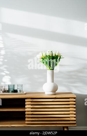 Skandinavische Innenausstattung mit Frühlingsstrauß aus weißen Tulpenblüten in Keramikvase, Tablett mit Kerzen auf einem Holzschrank. Minimalistisches Design Stockfoto
