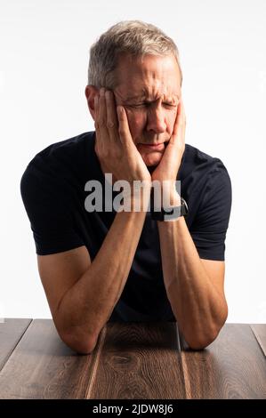 Älterer Mann in einem blauen T-Shirt, der seinen Kopf hält, auf einem dunklen Holztisch mit einem reinen, brillanten weißen Hintergrund, der schmerzt, gestresst und besorgt aussieht Stockfoto