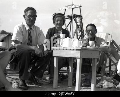 1930s, historisch, am Meer, Menschen sitzen neben einem kleinen Tisch mit Teekannen, mit einer Tasse Tee am Strand, Belgien. Auf dem Schild auf der Aussichtsplattform des Rettungsschwimmers steht: „Zee en Zonnebad Termes“ (Meer und Sonnenliegen verfügbar) Stockfoto