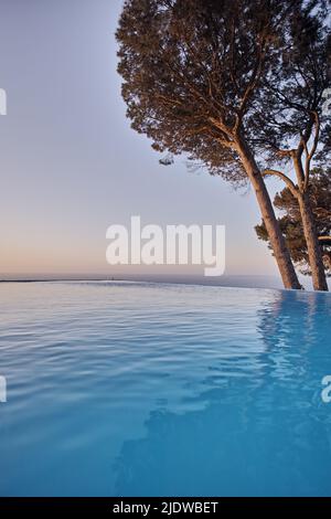 Ein unendlicher Pool mit Bäumen vor blauem Himmel mit Copyspace. Schöner Außenpool in einem Resort oder einer luxuriösen Ferienvilla Stockfoto