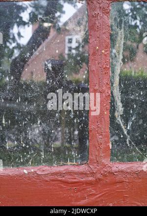 Nahaufnahme eines alten schmutzigen Fensters mit Blick auf die Nachbarschaft. Das Innere eines unsauberen Glasfensters mit roter, plumpiger Farbstruktur auf dem Rahmen Stockfoto