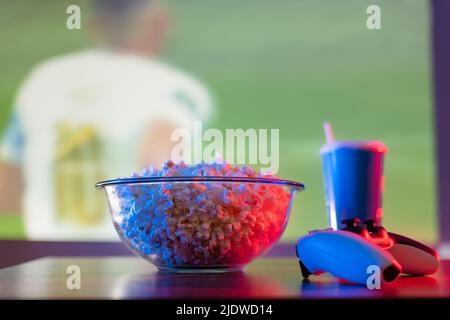 Vor dem Hintergrund eines Fußballspiels auf der großen Fernsehleinwand - Popcorn, ein Getränk und ein Joystick. Sportspiele, Online-Spiele mit Freunden, Sportspiele Stockfoto