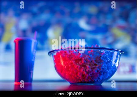 Vor dem Hintergrund eines großen Bildschirms eines Plasmafernsehens, einer Schüssel Popcorn, einem Getränk in einem Plastikglas auf einem Holztisch. Blaue Neonbeleuchtung. Ansehen Stockfoto