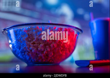 Nahaufnahme. Eine große Glasschüssel mit Popcorn und eine TV-Fernbedienung auf einem Holztisch vor dem Fernseher. Neonbeleuchtung. Abendruhe, Freizeit watc Stockfoto