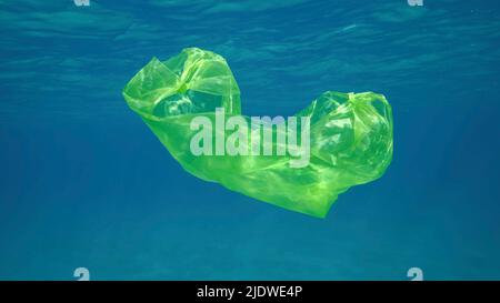 Plastikverschmutzung, Gelbe Plastiktüte schwimmt unter Wasser reflektiert auf der Oberfläche des blauen Wassers in Sonnenstrahlen. Weggeworfene Plastiktüten driften langsam Stockfoto