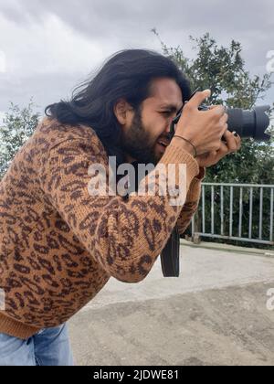 Nahaufnahme eines gut aussehenden jungen Mannes mit langen Haaren und Bart, der mit einer DSLR-Kamera (Digital Single-Lens Reflex) im Freien fotografiert Stockfoto