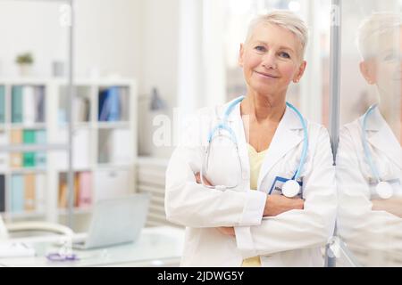 Content-Porträt Attraktive Ärztin mit kurzen blonden Haaren, die sich im eigenen Büro an einer glasigen Wand lehnt und Arme kreuzt Stockfoto