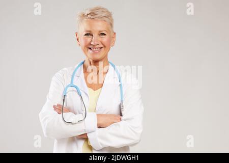 Porträt einer positiv reifen Hausärztin mit Stethoskop, das vor weißem Hintergrund steht und die Arme auf der Brust kreuzt Stockfoto
