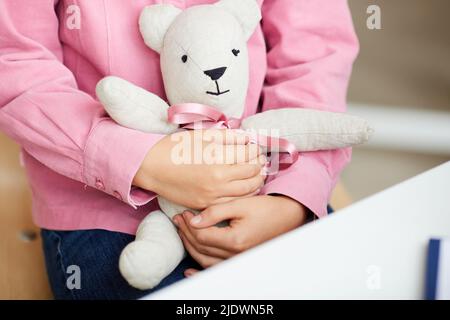 Nahaufnahme eines unerkennbaren Mädchens in rosa Hemd, das auf einem Stuhl sitzt und einen Spielzeugbären bei einem Arzttermin umarmt Stockfoto