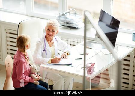 Lächelnder, freundlicher, reifer blonder Arzt mit Stethoskop am Hals, der im Büro am Schreibtisch sitzt und mit einem kleinen Patienten mit Spielzeug spricht Stockfoto