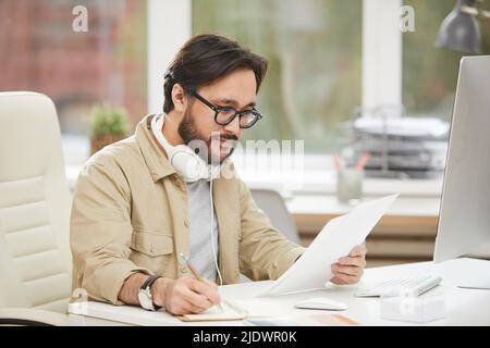 Inhalt hübscher junger asiatischer App-Designer in einer Brille, die am Tisch sitzt und Notizen auf dem Notizblock macht, während er Zeichnungen der Benutzeroberfläche ansieht Stockfoto