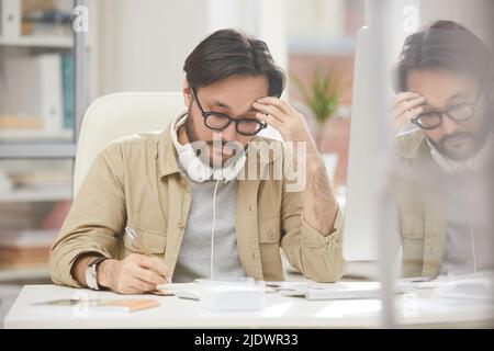 Rätselhafter Hipster junger asiatischer Mann mit Brille, der Kopfhörer am Hals trägt, sitzt am Tisch und liest Notizen im Tagebuch, während er an einem Projekt im Büro arbeitet Stockfoto