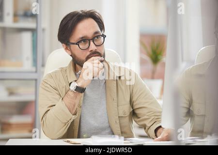 Serious pensive Hipster Asian Designer konzentriert sich auf Gedanken sitzen am Schreibtisch im Büro und Blick auf Monitor während der Analyse von Websites Stockfoto