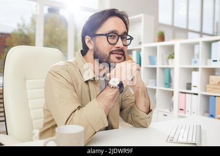 Content-Porträt ein verträumter junger asiatischer Designer mit Bart, der im Büro am Schreibtisch sitzt und sich mit dem Kopf auf den Händen lehnt Stockfoto