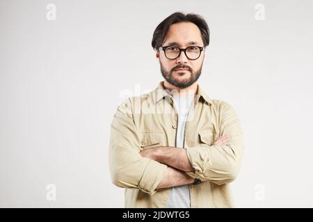 Porträt eines hübschen jungen asiatischen Mannes im Khaki-Hemd mit stilvoller Brille, die die Arme vor grauem Hintergrund kreuzt Stockfoto