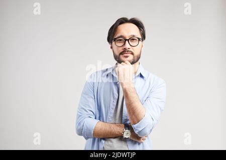 Porträt des Inhalts unternehmungslustiger junger asiatischer Mann mit Bart in lässigem Hemd und Brillen, die das Kinn in Gedanken berühren Stockfoto
