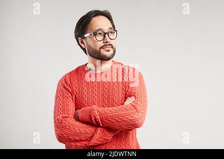Inhalt ein verträumter junger asiatischer Mann mit kühlem Bart und Schnurrbart trägt einen warmen Strickpullover, der die Arme auf der Brust kreuzt Stockfoto