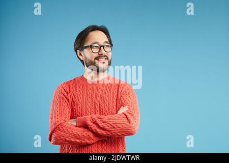 Fröhlicher, verträumter junger asiatischer Mann mit Bart und Schnurrbart, der einen stylischen Pullover auf blauem Hintergrund trägt und dabei verträumt davonschaut Stockfoto