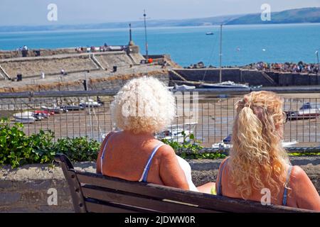 Rückansicht von zwei Frauen, die in der Sommersonne auf einer Parkbank sitzen und den Pier und Hafen von New Quay in Ceredigion West Wales, Großbritannien, BETRACHTEN KATHY DEWITT Stockfoto