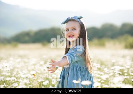 Lächelnd nettes Baby Mädchen 3-4 Jahre alt tragen stilvolle Kleid mit Blumen in blühenden Kamille Wiese im Freien. Kindheit. Sommersaison. Stockfoto