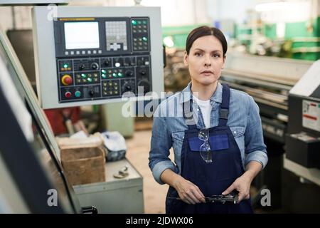 Porträt einer attraktiven Fabrikarbeiterin in Overalls, die an einer modernen Industriemaschine steht und Mikrometer hält Stockfoto