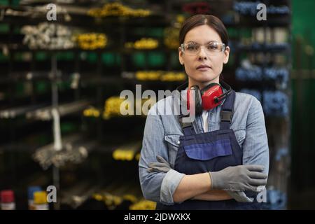 Seriöse, selbstbewusste, professionelle Arbeiterin der Fabrik, die Handschuhe und Overalls trägt und im Lager der Metallverarbeitung steht und die Kamera anschaut Stockfoto