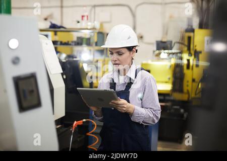 Konzentrierte junge CNC-Maschinenbedienerin in Hardhat, die an der industriellen Drehbank steht und die Setups auf einem digitalen Tablet überprüft Stockfoto