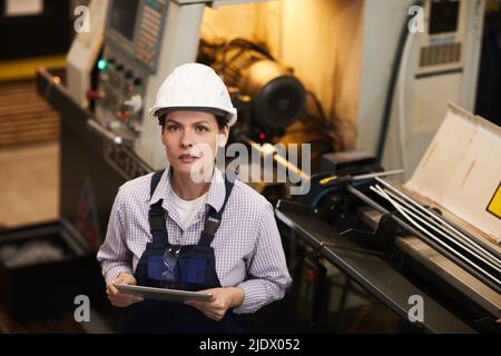 Content-Porträt Attraktive Frau in weißem Hardhat an der Drehbank stehend und mit Tablet, sie arbeitet in der Fertigungsindustrie Stockfoto